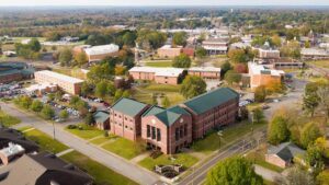 Aerial view of a college campus with several red-brick buildings, tree-lined streets, parked cars, and a small town in the background surrounded by green and autumn-colored trees.