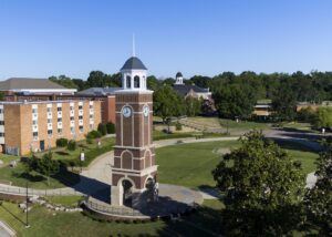 A tall brick clock tower stands in a landscaped grassy area with sidewalks, surrounded by trees and campus buildings under a clear blue sky.