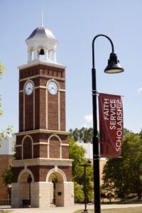 A brick clock tower with a white roof stands under a blue sky. In the foreground, a lamppost displays a red banner reading FAITH SERVICE SCHOLARSHIP. Green trees and campus buildings surround the scene.
