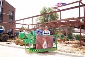 Two people in hard hats and safety gear stand on a green lift holding a banner with a white dome logo, at a construction site with steel framework and an American flag above them.