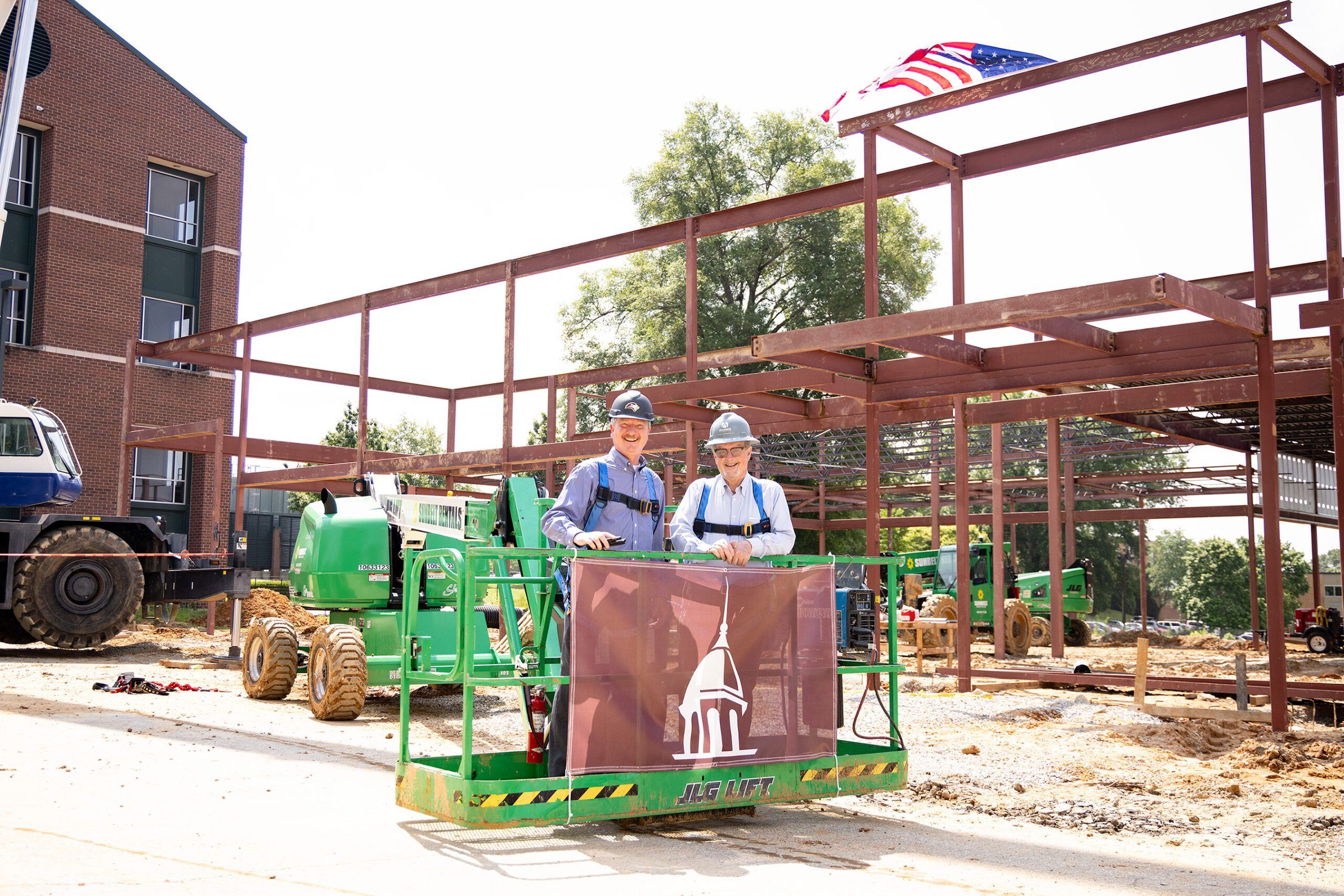 Two people in hard hats and safety gear stand on a green lift holding a banner with a white dome logo, at a construction site with steel framework and an American flag above them.