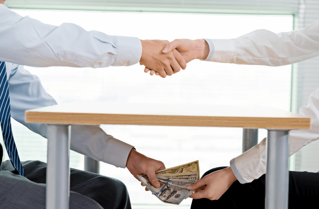 Two people in business attire shake hands over a table while exchanging cash under the table, suggesting a secretive or corrupt financial deal.