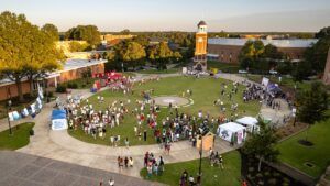 A large group of people gather on a grassy campus quad with tents and booths set up; a clock tower and several buildings are visible in the background on a sunny day.
