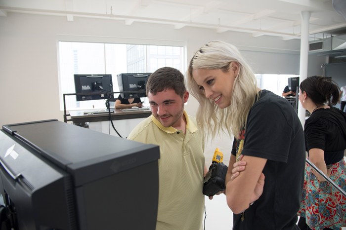 A man in a yellow shirt and a woman with long blonde hair smile while looking at a computer monitor in a bright, modern office space. Other people are working in the background.