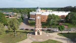 A red brick clock tower stands in the center of a circular plaza on a grassy campus, surrounded by trees, parked cars, and academic buildings under a clear sky.