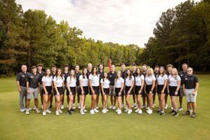 A group of young men and women, wearing matching golf uniforms, stand together on a golf course green, with two adult coaches at each end. Trees and a flag are visible in the background under a partly cloudy sky.