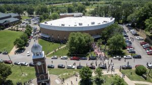 Aerial view of a large crowd gathered outside a circular building with a clock tower nearby, surrounded by trees, parked cars, and open grassy areas on a sunny day.