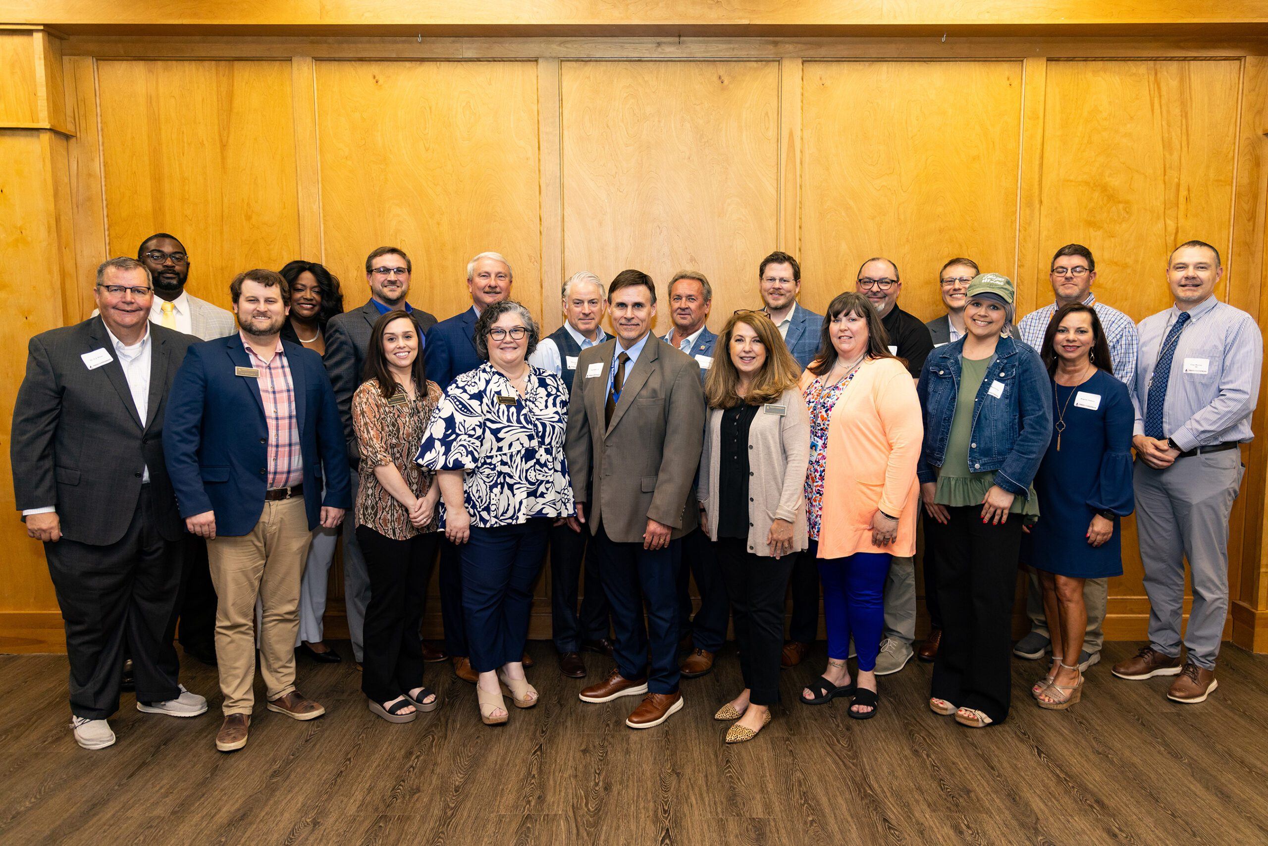A group of 21 adults, both men and women, stand posing and smiling for a group photo indoors against a wooden wall. Most are dressed in business or business-casual attire and are wearing name tags.