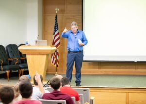 A man in a blue shirt speaks and gestures while standing at the front of a lecture hall, with an American flag, podium, and projection screen behind him; audience members sit facing him.