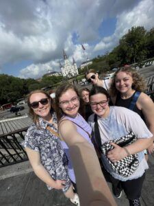 A group of six smiling young people take a selfie outdoors near a black fence, with a historic white cathedral and cloudy sky in the background. Trees and parked cars are visible behind them.
