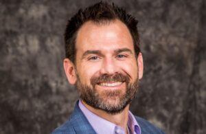 A man with short dark hair and a beard smiles at the camera. He is wearing a blue checked blazer over a light purple shirt. The background is mottled gray.