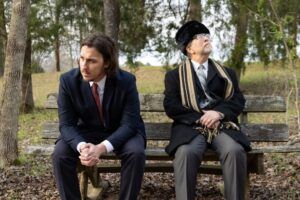 Two men in suits sit on a wooden bench outdoors. The younger man looks down with hands clasped, while the older man in a hat and striped scarf gazes upward. Trees and fallen leaves surround them.