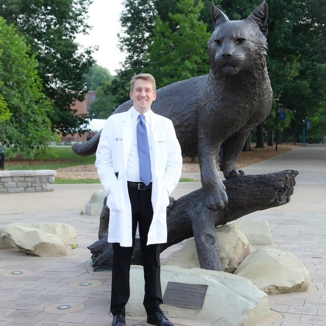 A man in a white lab coat stands smiling in front of a large wildcat statue on a paved area, with trees and a campus-like setting in the background.