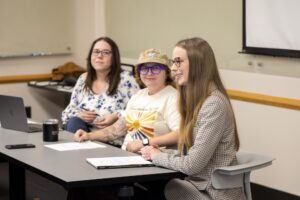 Three women sit at a conference table in a classroom setting. One speaks while the others listen. Papers, a laptop, and a tumbler are on the table. A whiteboard and projector screen are visible in the background.