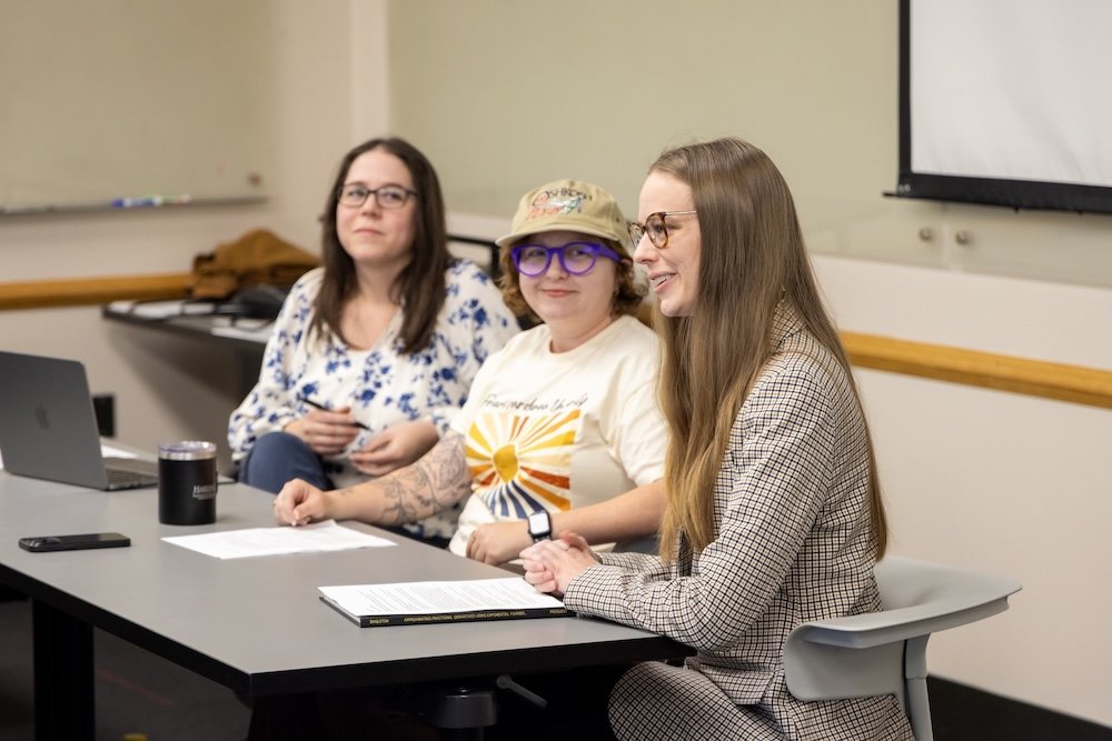 Three women sit at a conference table in a classroom setting. One speaks while the others listen. Papers, a laptop, and a tumbler are on the table. A whiteboard and projector screen are visible in the background.