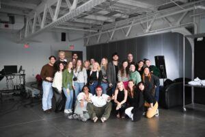 A group of 22 people pose together in a brightly lit, industrial-style studio with high ceilings, concrete floors, and photo equipment in the background. Most are smiling, and two people sit cross-legged at the front.