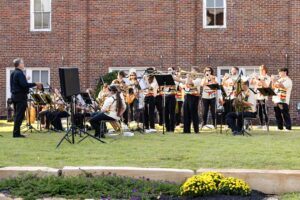 A marching band in matching uniforms plays brass and woodwind instruments outside on a lawn, with a conductor leading them. A brick building is in the background.