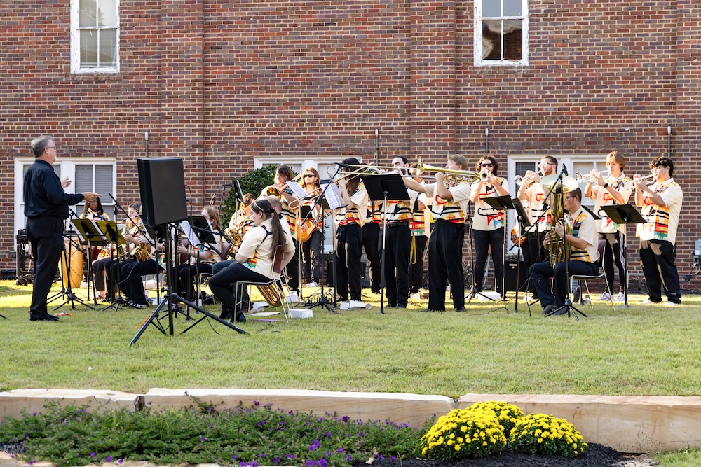 A marching band in matching uniforms plays brass and woodwind instruments outside on a lawn, with a conductor leading them. A brick building is in the background.