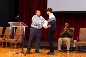 A man shakes hands with a young man receiving an award on stage while another man sits nearby, clapping. Three empty chairs and a podium are also visible, with a red curtain in the background.
