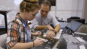 A man and a woman work together on an electronics project at a table. The woman is assembling components on a board while the man points and offers guidance. A laptop and small containers with parts are nearby.