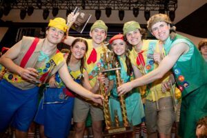 A group of six young adults in colorful scout-themed costumes smile and hold a large trophy together, celebrating on stage under bright lights.