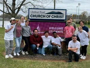 A group of twelve smiling people pose together in front of a Church of Christ sign that says Welcome Students. Most are casually dressed, and they stand on grass with trees and a sports field in the background.
