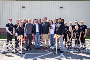 A group of people, including a women’s softball team in black uniforms and several adults in casual attire, stand smiling behind a red ribbon outside a building on a sunny day.