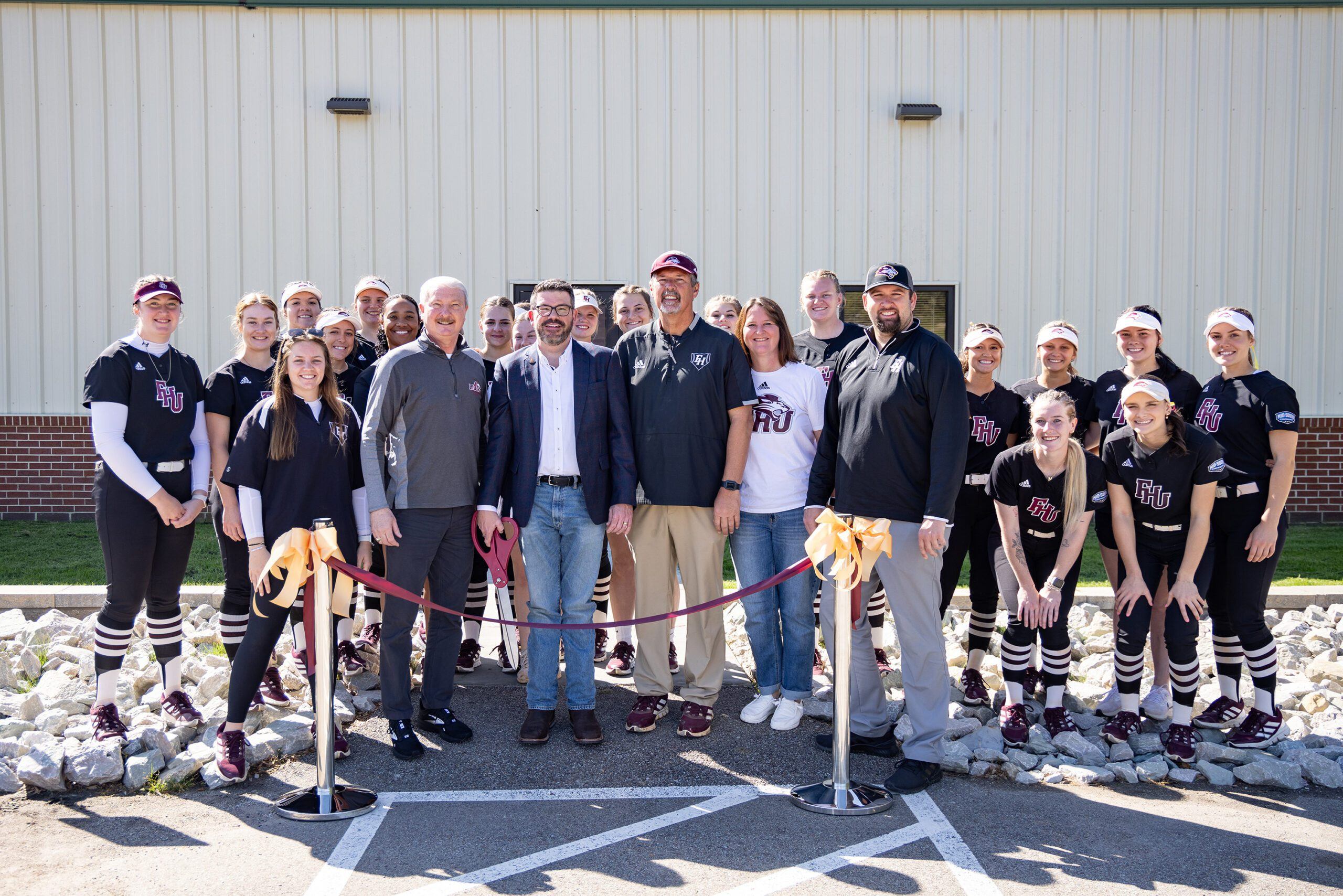 A group of people, including a women’s softball team in black uniforms and several adults in casual attire, stand smiling behind a red ribbon outside a building on a sunny day.