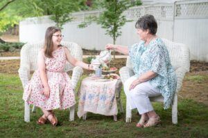 A young girl and an older woman sit on white wicker chairs outdoors, smiling as the woman pours tea. A small table between them holds a teapot, cups, and treats. Both are dressed in light, floral-patterned clothing.