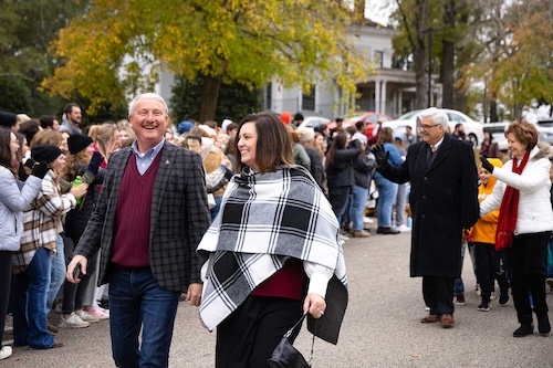 A smiling man and woman walk outdoors among a crowd of people, many of whom are waving. The group is surrounded by autumn trees and houses in the background.