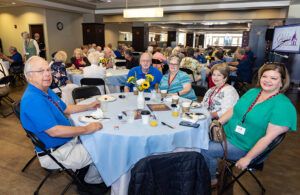 A group of older adults sit around a round table with light blue tablecloths, smiling at the camera. The room is full of people and decorated with sunflowers. Food, drinks, and name tags are visible on the table.