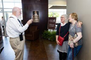 An older man takes a photo of two smiling elderly women who are standing arm-in-arm in a lobby area. A welcome sign and a plaque are visible in the background.