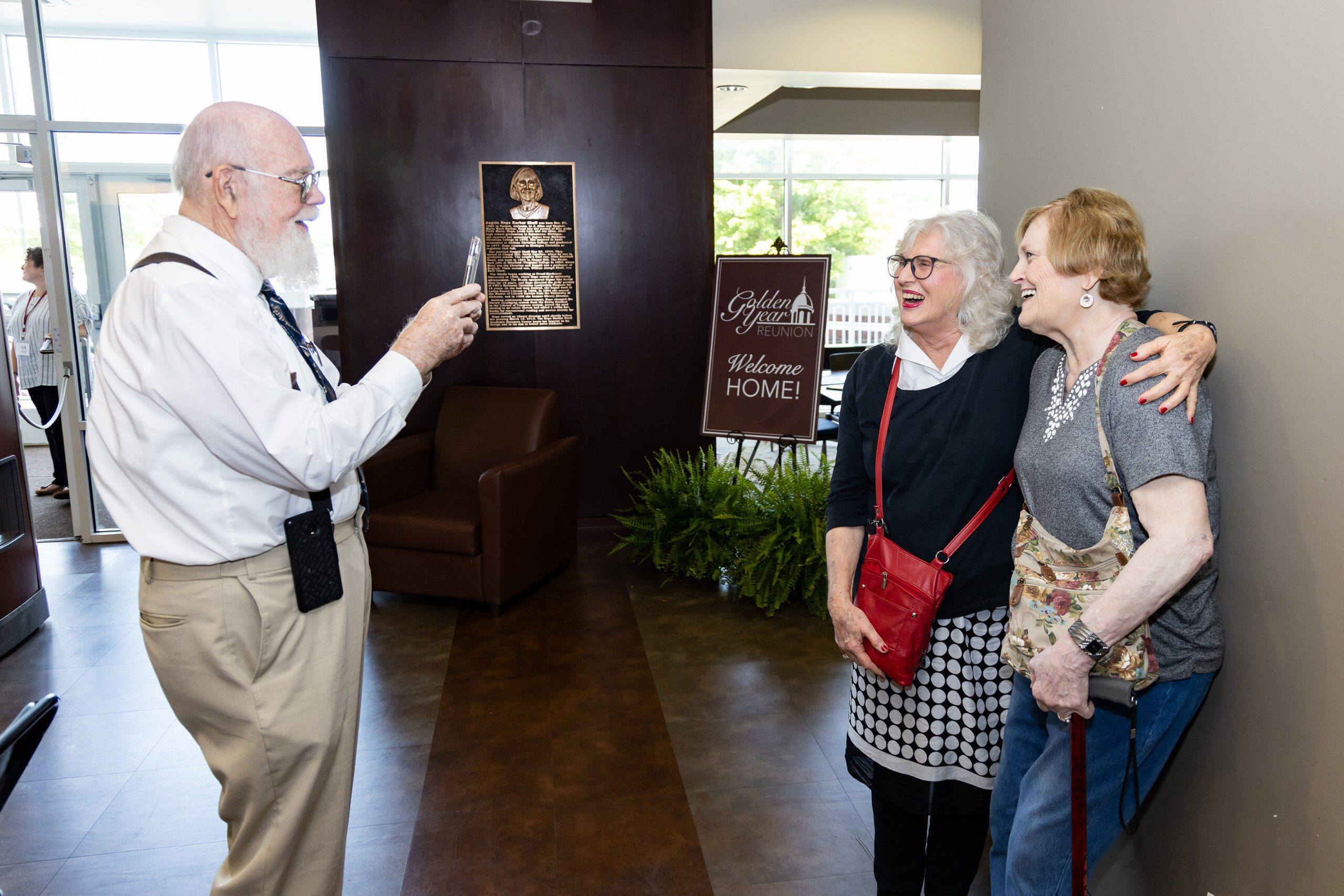 An older man takes a photo of two smiling elderly women who are standing arm-in-arm in a lobby area. A welcome sign and a plaque are visible in the background.