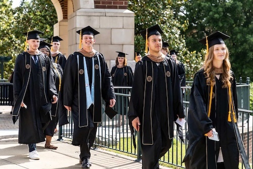 A group of smiling graduates in caps and gowns walk outdoors on a sunny day, near a building with brick and stone columns and green trees in the background.