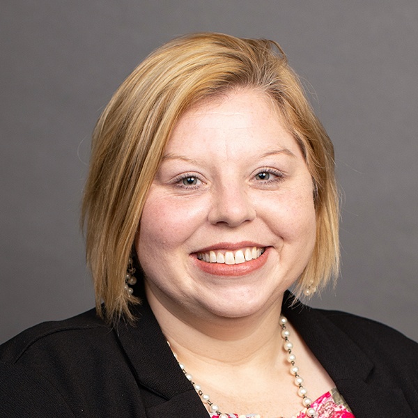 A woman with short blonde hair smiles at the camera. She is wearing a black blazer, a pink floral top, a pearl necklace, and pearl earrings. The background is plain and gray.