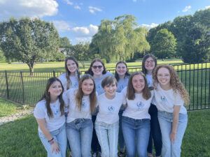 A group of ten young women stand close together outside on grass, all smiling and wearing matching white T-shirts and jeans. Behind them is a black fence, green trees, and a blue sky with clouds.