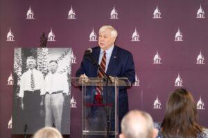 An older man in a suit speaks at a podium with a microphone. Behind him is a maroon backdrop with white building icons and a large black-and-white photo of two men. Audience members are visible in the foreground.