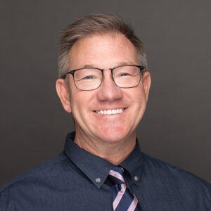 Smiling man with short light brown hair, wearing glasses, a dark button-up shirt, and a striped tie, posing in front of a plain gray background.