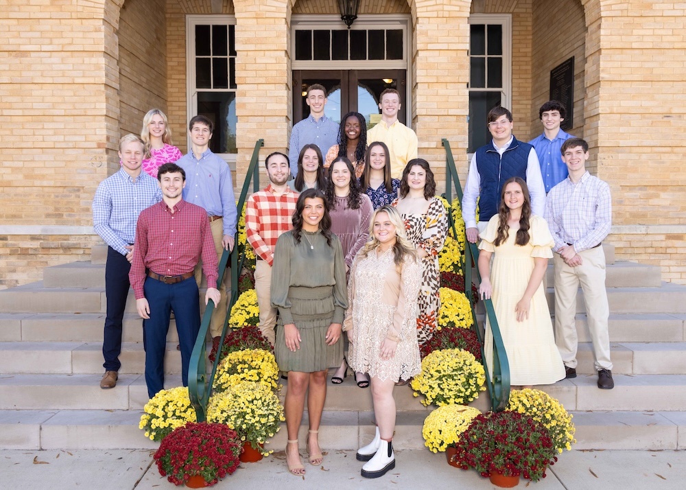 A group of seventeen young adults, dressed in semi-formal attire, pose on steps decorated with yellow, white, and red chrysanthemums in front of a brick building with large windows and double doors.