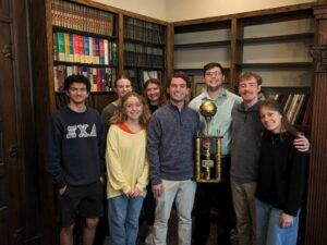 Seven smiling young adults stand together in a cozy library with wooden shelves and books. One person holds a large trophy with a gold basketball on top. They appear happy and celebratory.