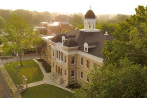 Aerial view of a historic brick building with a white cupola and arched windows, surrounded by trees and greenery, with a soft, golden sunrise in the background.