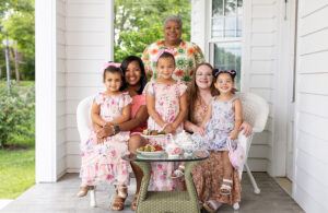 Four smiling women and girls pose together on a white porch. Three young girls in floral dresses sit in front, with two women seated behind them and an older woman standing. A tea set and snacks are on the glass table.