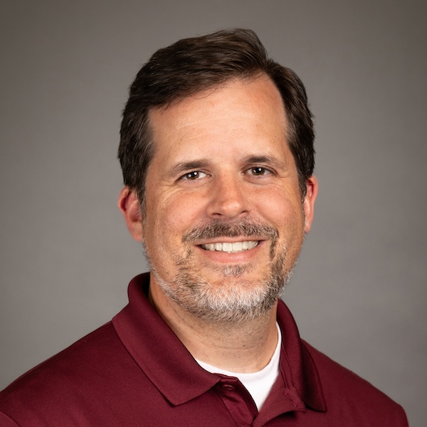A man with short brown hair, a beard, and mustache is smiling. He is wearing a light blue collared shirt and a red bow tie with a beige center, posed in front of a plain gray background.
