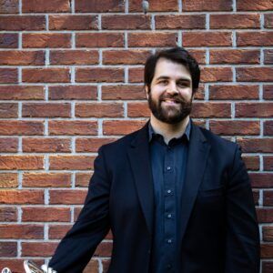 A man with dark hair and a beard, wearing a black suit and shirt, stands smiling in front of a brick wall. He is holding a silver musical instrument, partially visible on the left side of the image.