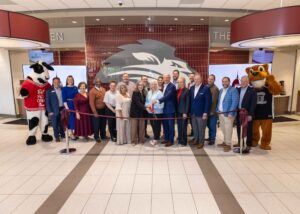 A group of people pose indoors for a ribbon-cutting ceremony in front of a lion mural, flanked by two mascots—a cow in a red shirt and a lion in a jersey. The group is smiling and holding large scissors.