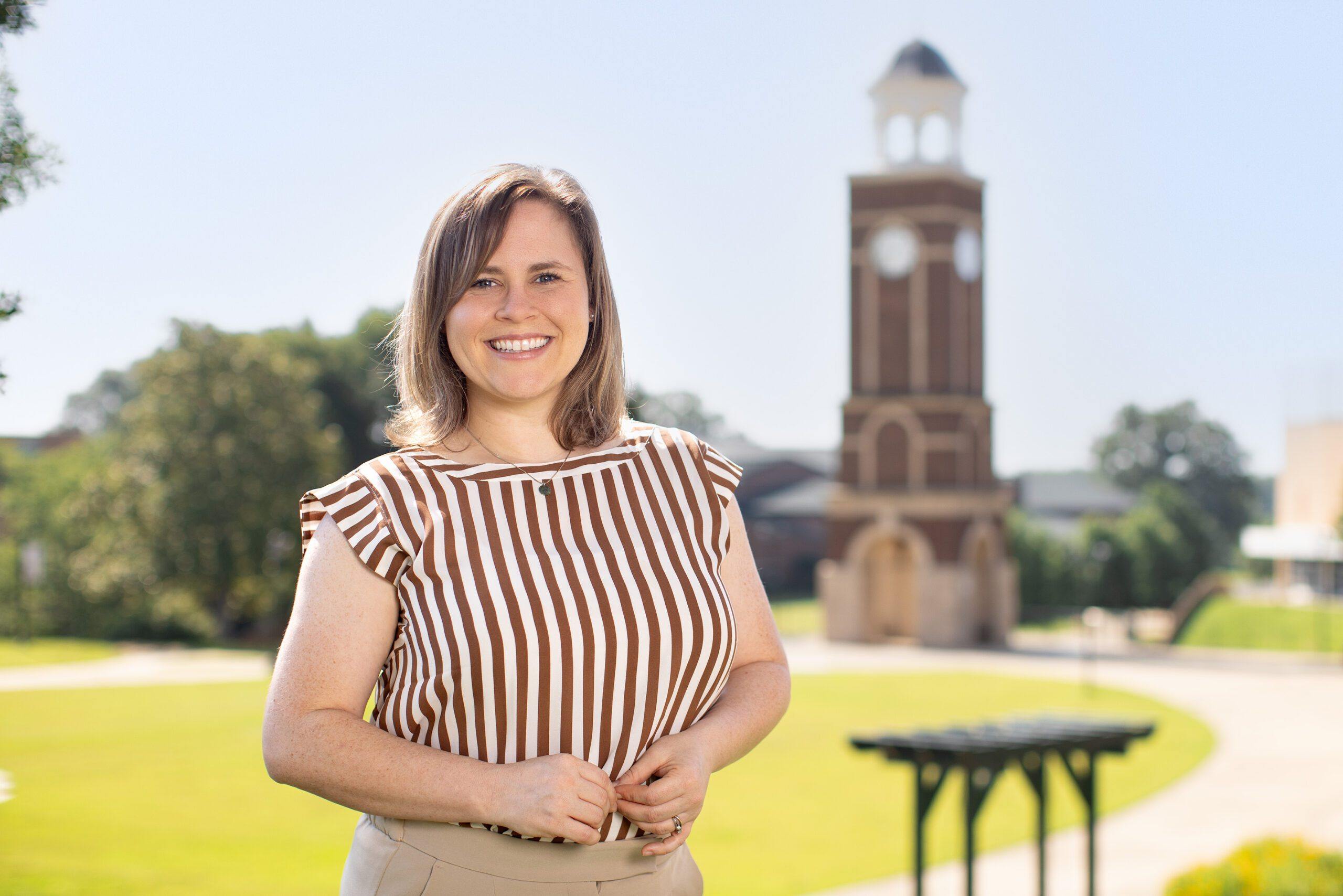 A woman with shoulder-length brown hair, wearing a brown and white striped blouse, smiles while standing outdoors in front of a brick clock tower on a sunny day.