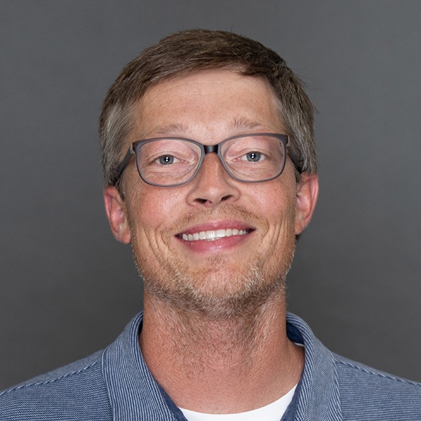 A man with short brown hair, light stubble, and glasses smiles at the camera. He is wearing a blue collared shirt and is posed in front of a plain gray background.