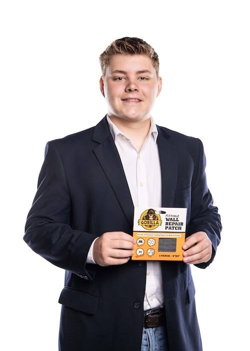A young man in a navy blazer and white shirt smiles while holding a package of Gorilla Waterproof Patch & Seal Tape against a white background.