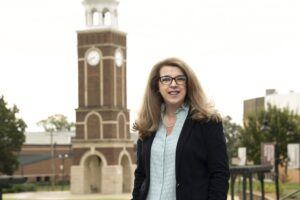 A woman with long, light brown hair and glasses stands outdoors, smiling, wearing a blue blouse and dark blazer. A tall brick clock tower is visible in the background. Trees and buildings surround the area.