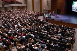 A large audience sits in a spacious auditorium, facing a stage where a speaker stands at a podium. Four empty chairs and a screen displaying an image are also on the stage. Most seats are occupied.